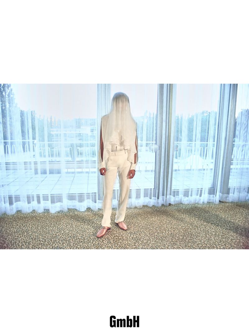 Model wearing tailored off-white trousers and a sheer white veil covering the head, standing in front of sheer curtains.