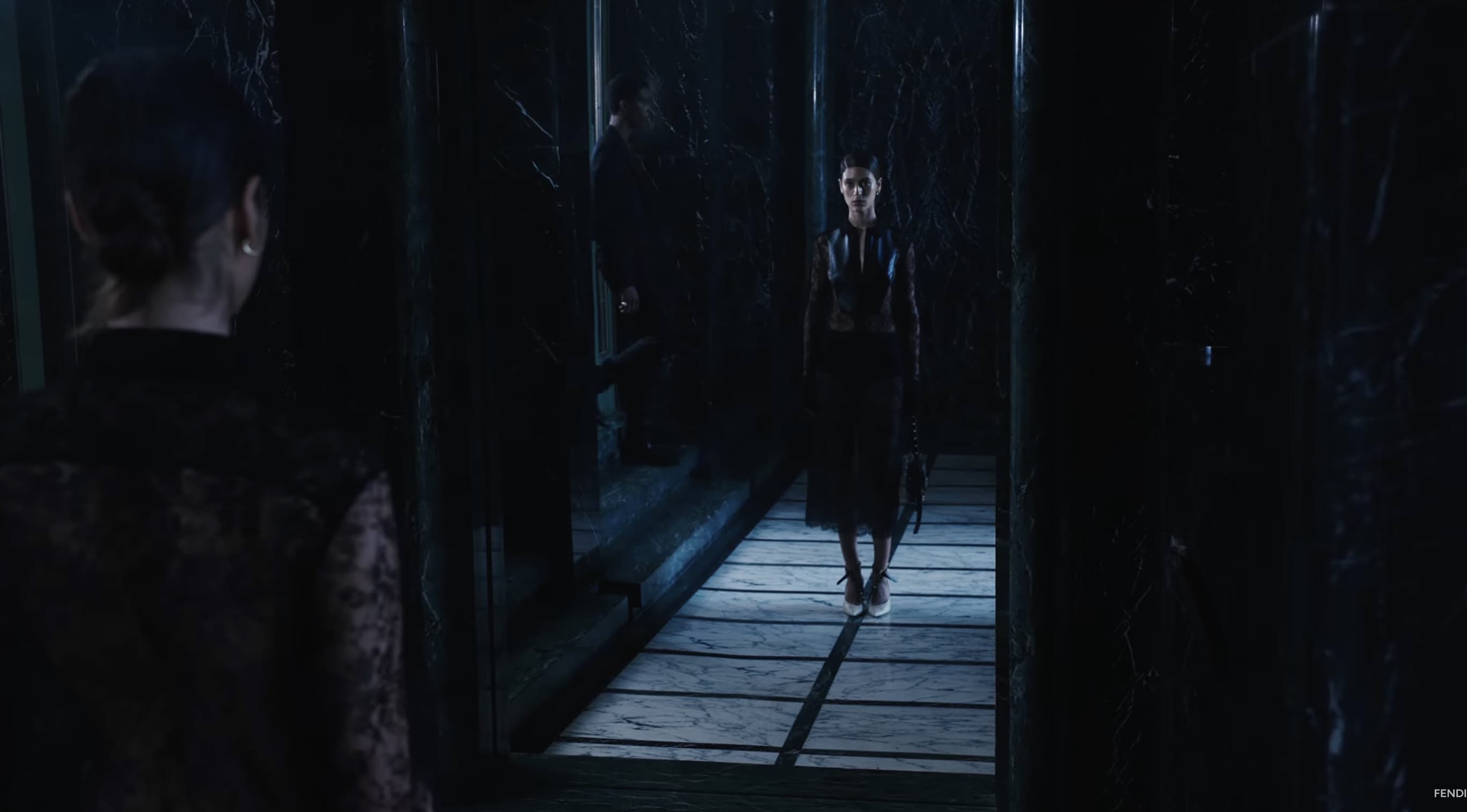 Fashion shoot in a dark hallway with a woman in a black outfit posing as the primary subject, alongside a second woman in the foreground.