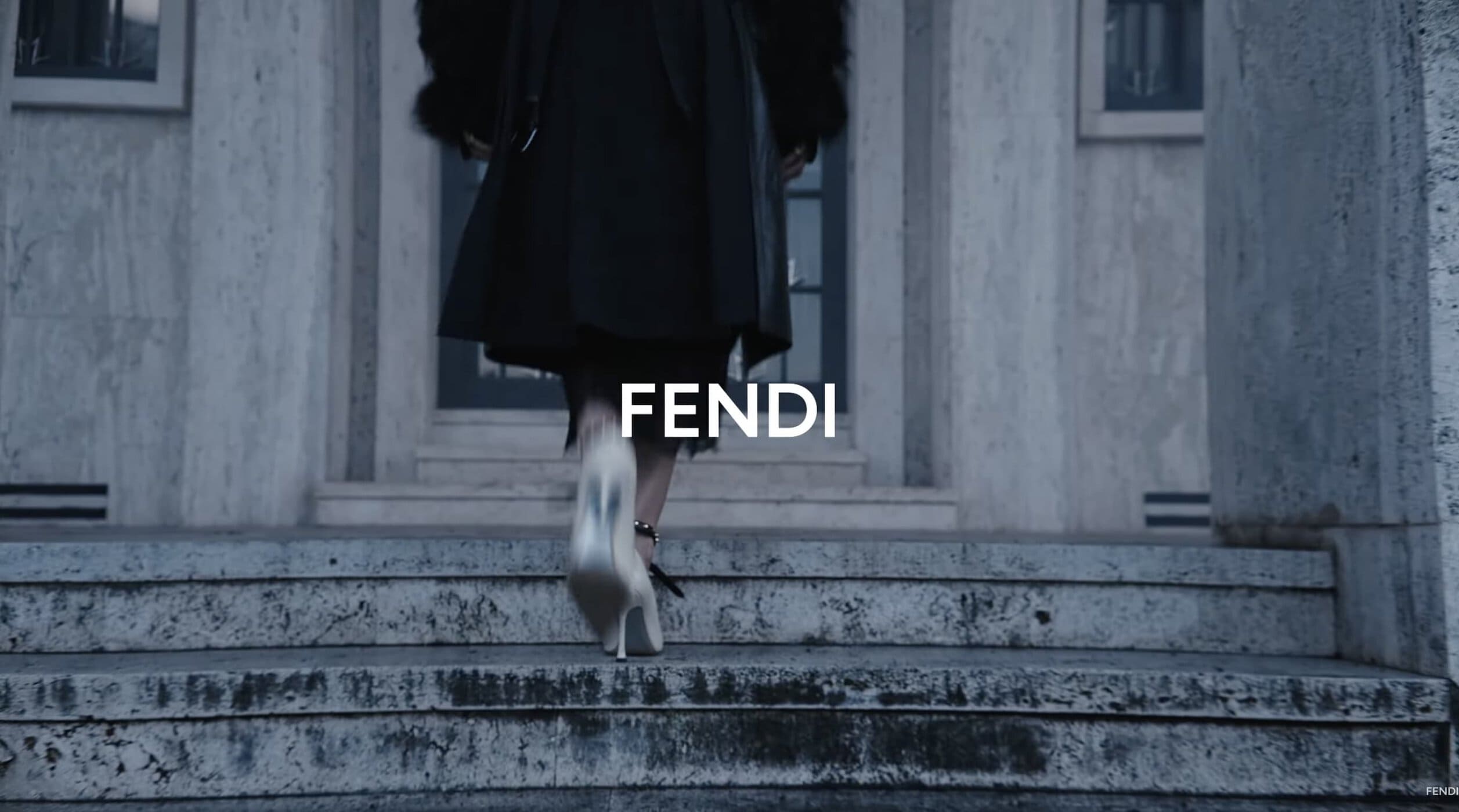 Person in a black coat and light stiletto heels walking up weathered stone stairs with white Fendi typography overlay.