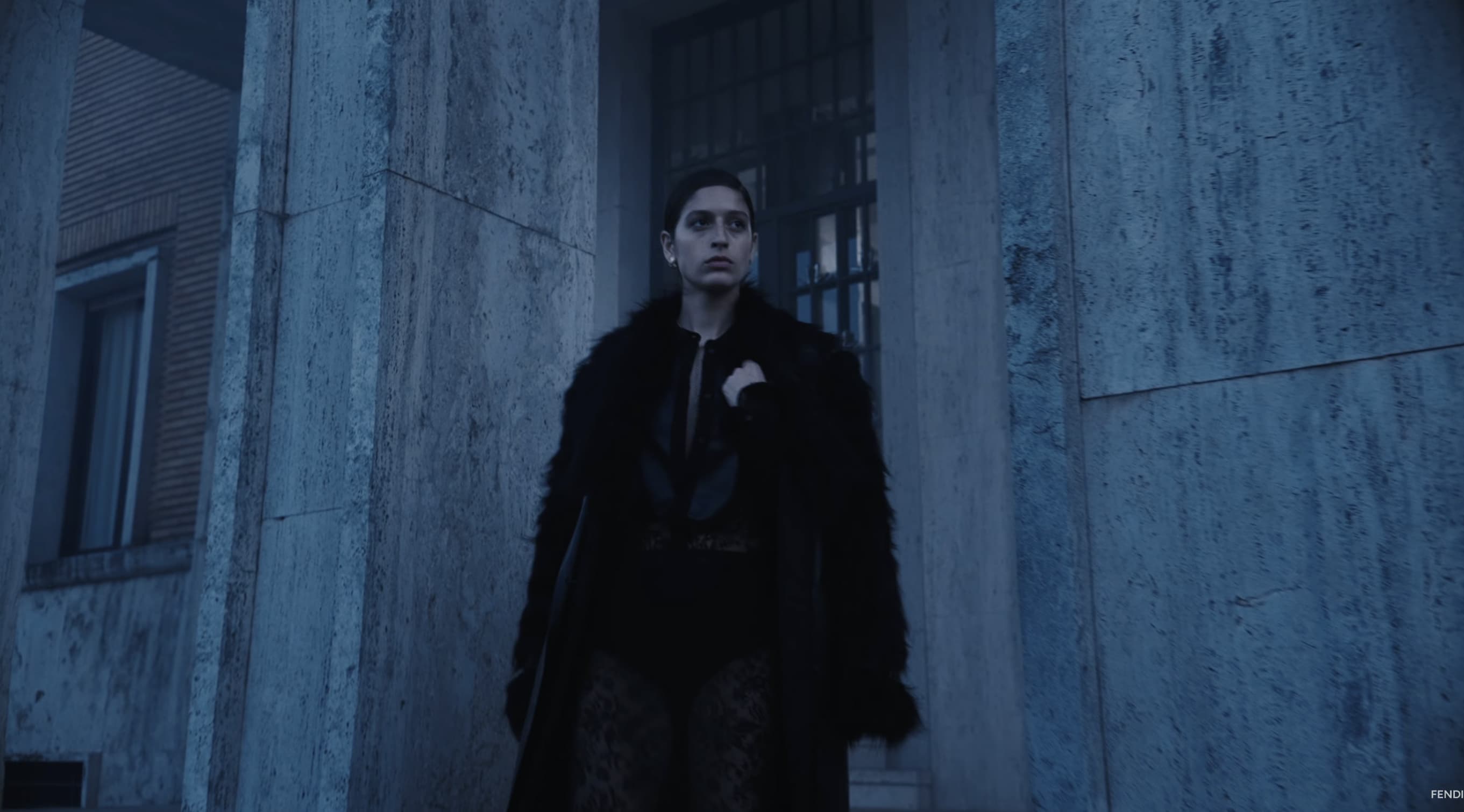 Model in a black textured fur coat stands between stone architectural columns under cool, blue-toned atmospheric lighting.