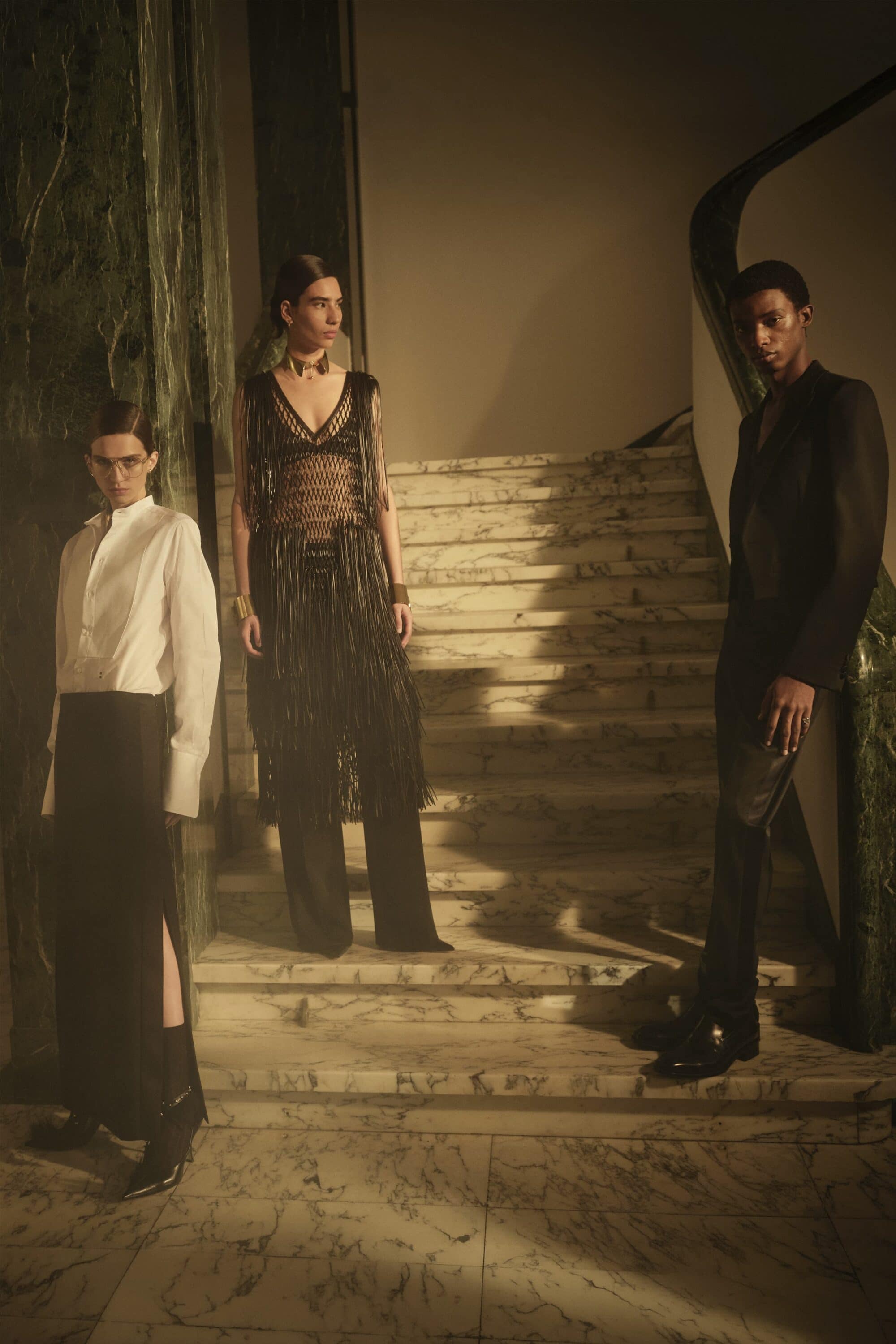 Three fashion models in elegant black and white tailored eveningwear posing on an opulent marble staircase with dark green marble accents.