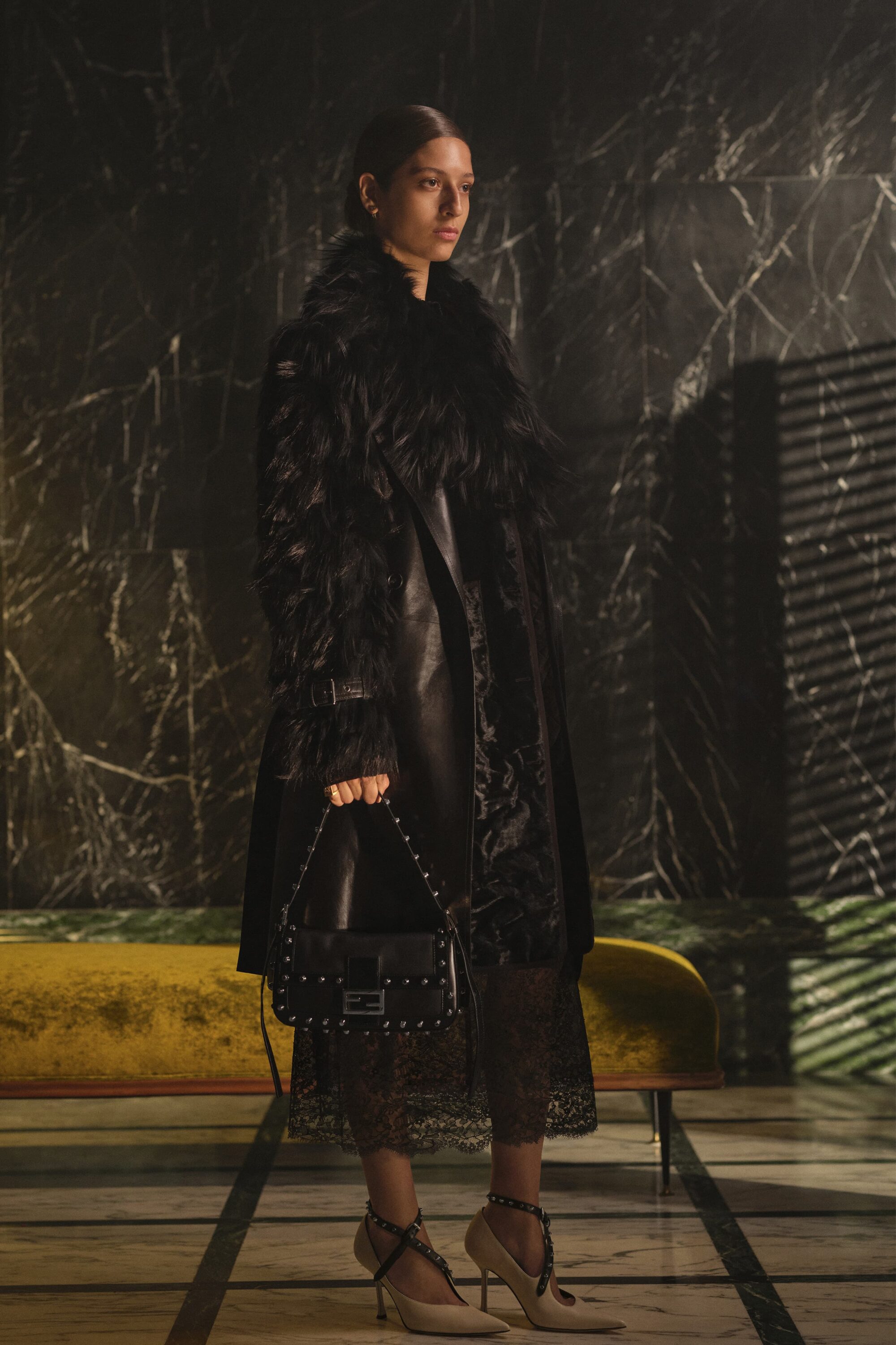 Model in a black feather coat holding a studded black handbag in front of a dark marble wall.