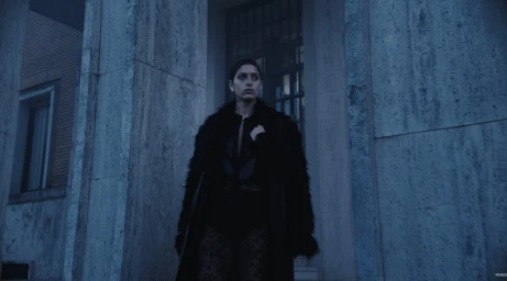 Model in a black textured fur coat stands between stone architectural columns under cool, blue-toned atmospheric lighting.