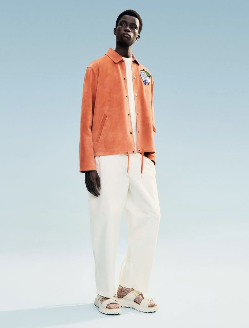A full-length shot of a Black man standing against a light blue backdrop, wearing a light orange jacket, white pants, and sandals.
