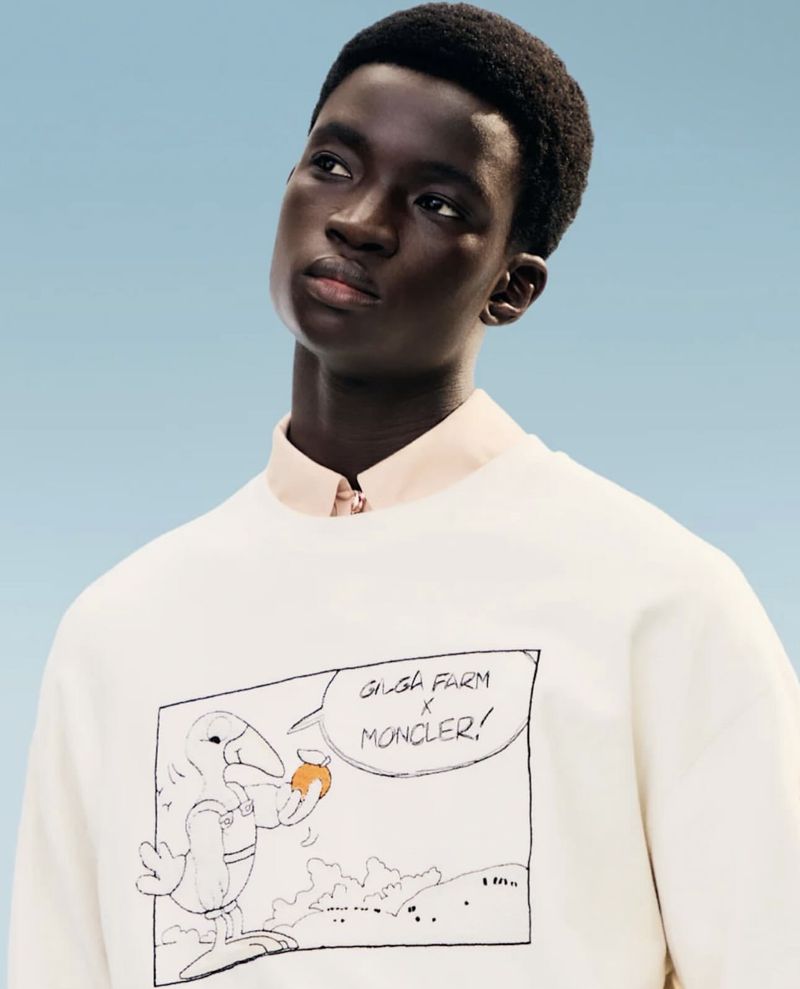 Adult male poses indoors wearing a crewneck sweatshirt with cartoon graphic, in a studio shot against a pale blue backdrop.
