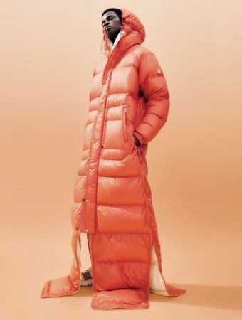 An adult male in a puffy, long, bright-orange down coat stands against a light-orange backdrop in a fashion studio, creating a modern, editorial look.