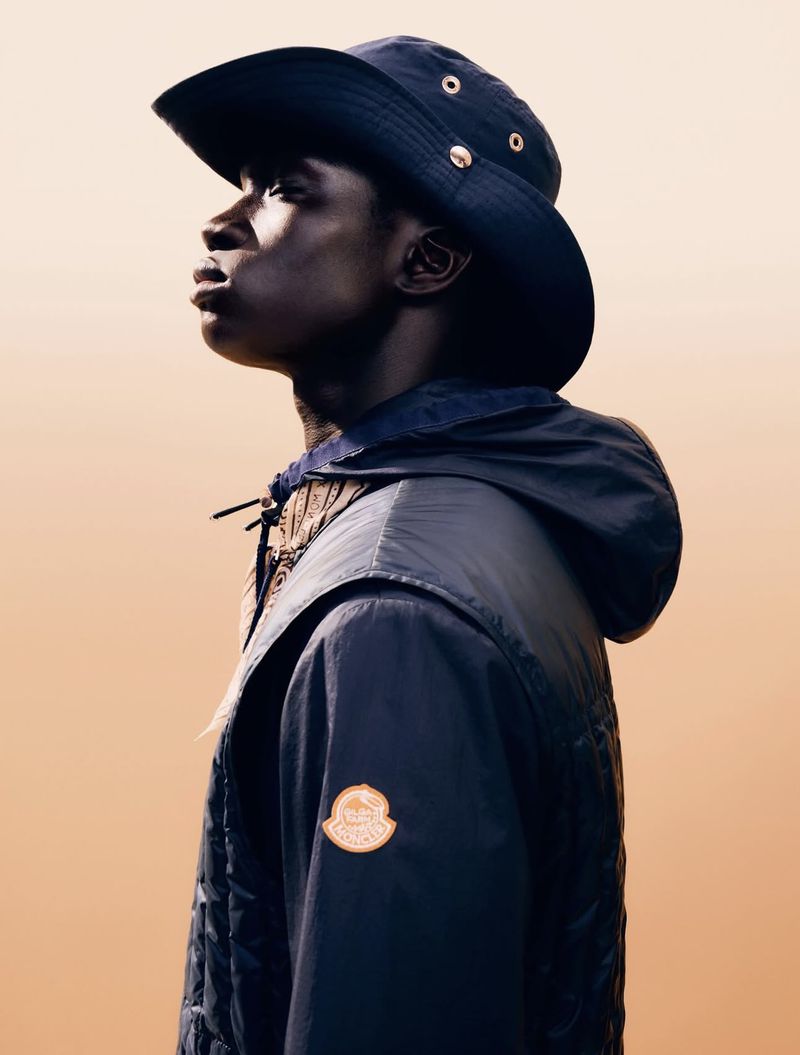 Black male model in profile wearing a dark navy wide-brim hat and hooded Moncler jacket against a warm gradient background.