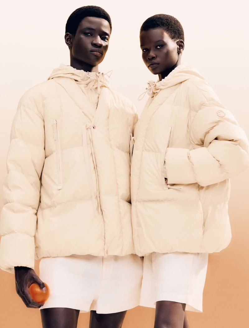 Two models wearing cream-colored hooded puffer jackets and white shorts pose in a studio setting with a cream to tan gradient backdrop.