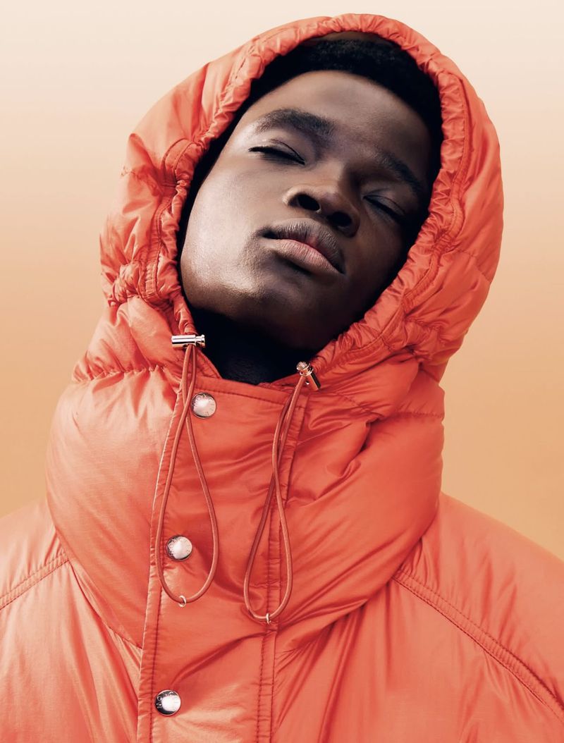 Portrait of a Black adult male in an orange puffer jacket with a hood, eyes closed, against a light-orange gradient background.