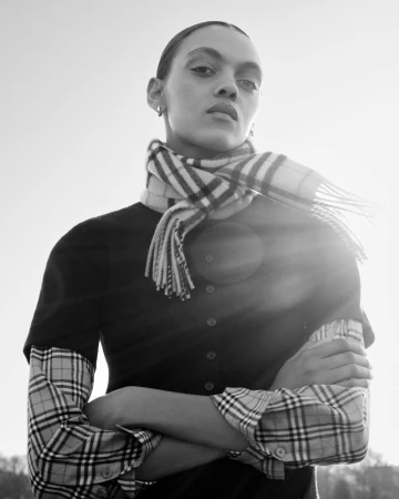Monochrome portrait of a model in a Burberry check scarf and layered check shirt with arms crossed outdoors in bright backlight.