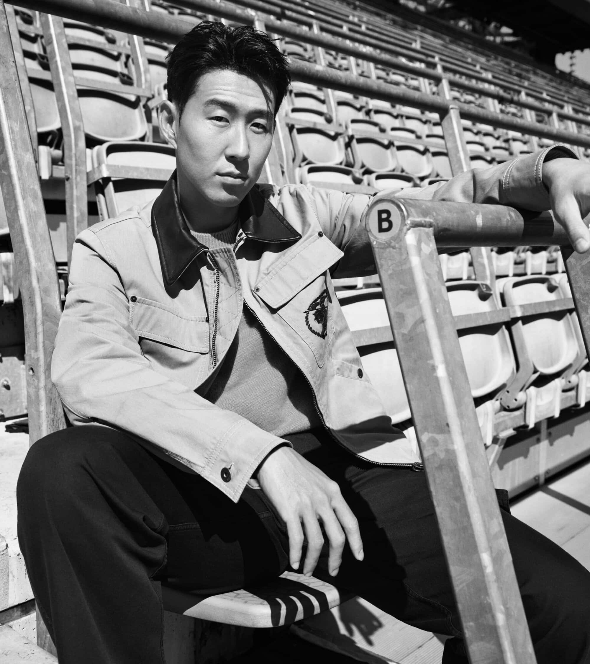 Son Heung-min sits in empty stadium bleachers wearing a collared utility jacket and dark trousers in a black and white portrait.