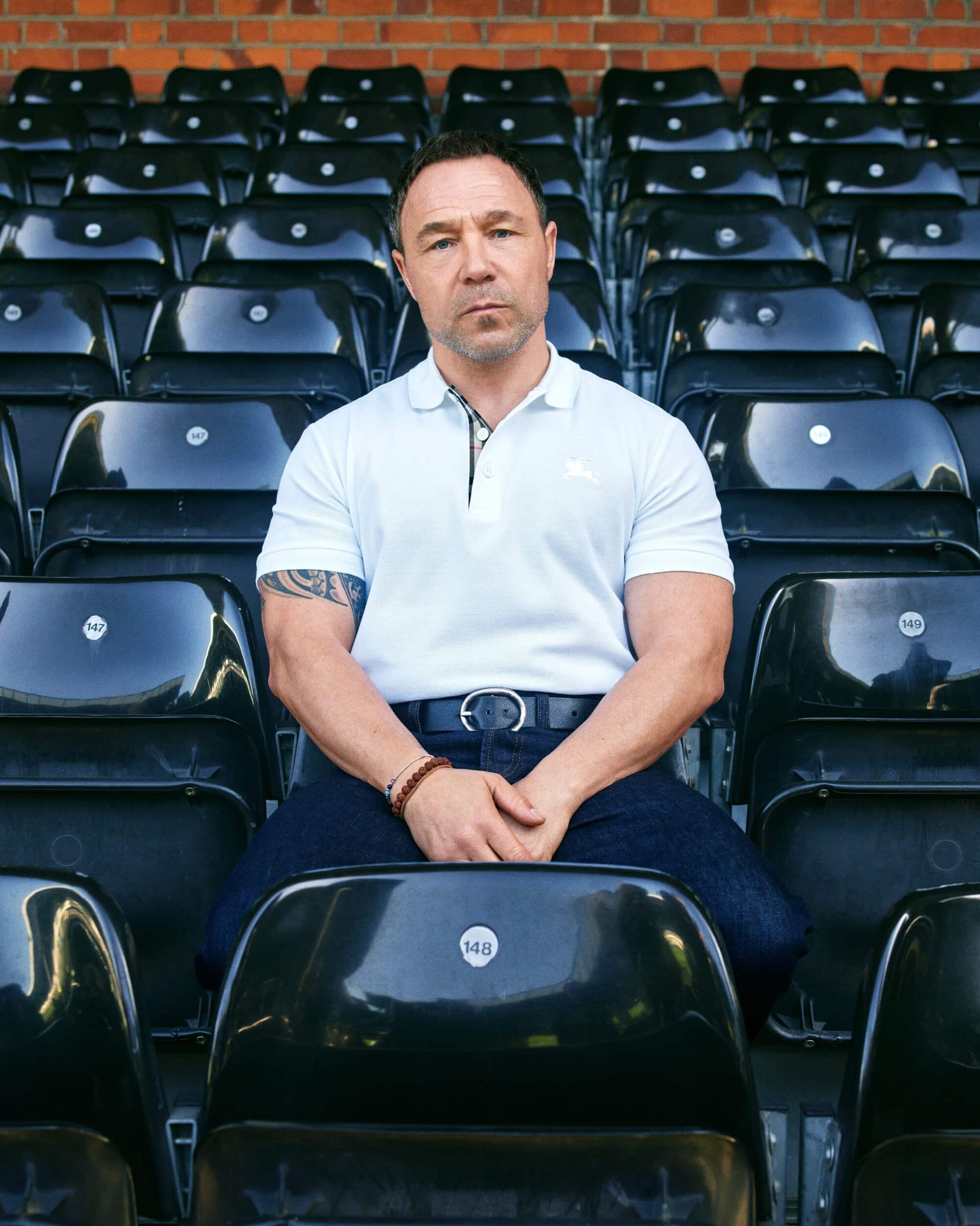 Stephen Graham seated in rows of empty stadium seats wearing a white polo shirt and dark trousers for Burberry Fall 2026 Ad Campaign