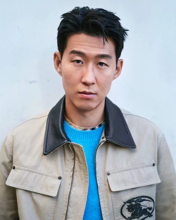 Footballer Son Heung-min wearing a beige collared jacket and bright blue knit sweater against a plain neutral backdrop.
