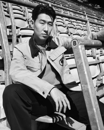 Son Heung-min wearing a collared utility jacket and dark trousers while seated on stadium bleachers in black and white.