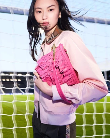East Asian woman holding a pink handbag wearing a light pink jacket against a soccer net background