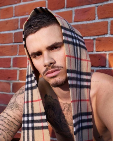 Romeo Beckham shirtless with a beige Burberry check scarf draped over his head against a red brick wall.