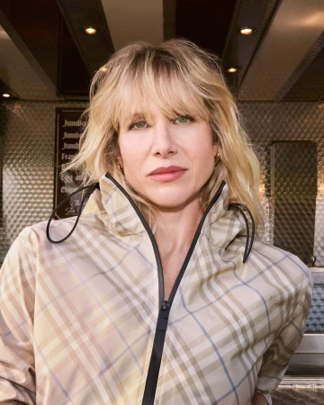 Lucy Punch looking forward wearing a beige Burberry check zip jacket with a high collar outside a shop window.