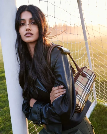 Neelam Gill in a black leather jacket with a Burberry check handbag leaning against a soccer goalpost.