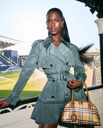 Jodie Turner-Smith poses in a belted teal trench dress holding a classic Burberry check handbag at a sports stadium.