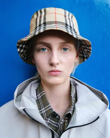 Model wearing a classic check bucket hat and off-white hooded jacket against a solid blue background.