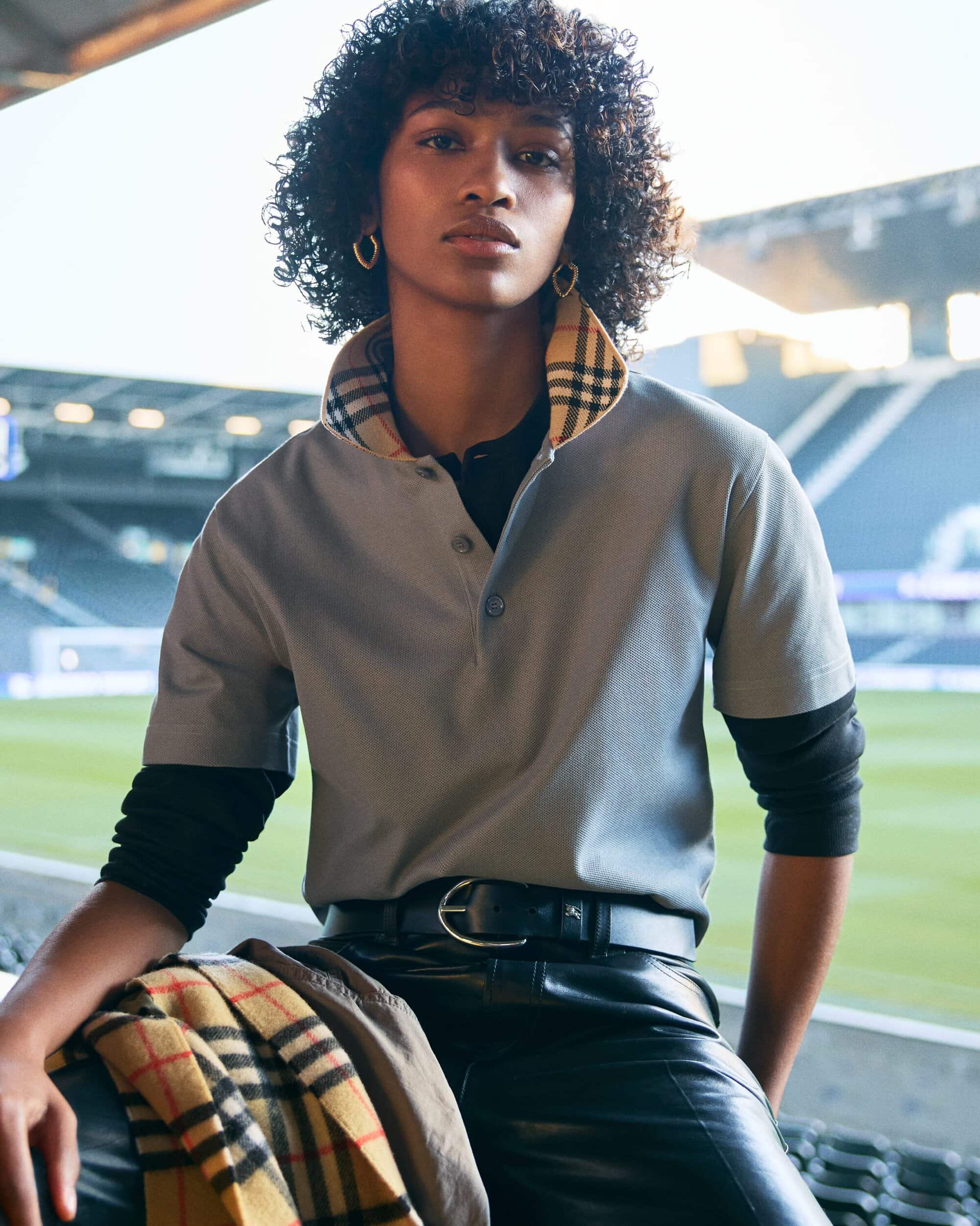 Model with curly hair in a grey polo shirt with check collar and black leather trousers seated in a sports stadium.