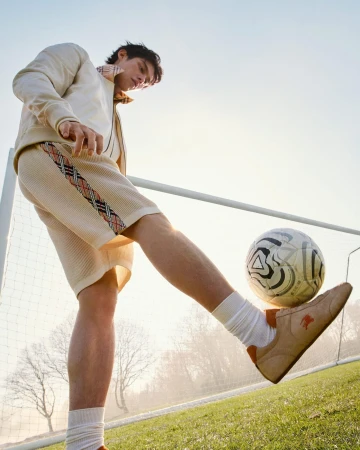Young man kicking a soccer ball into the air on an outdoor grass field in front of a goalpost, wearing a beige jacket and shorts.