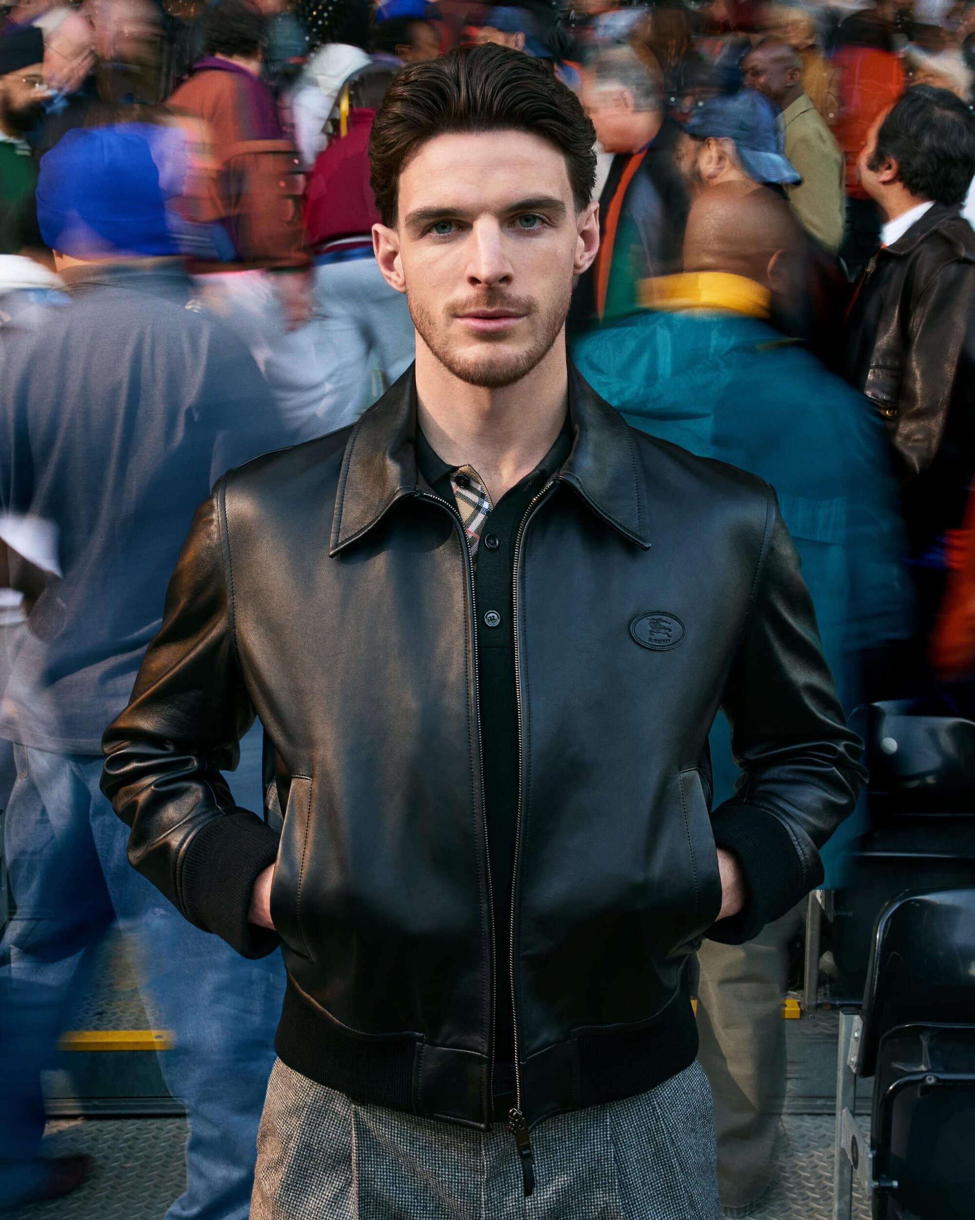 Declan Rice standing in a black leather jacket and grey trousers surrounded by a motion-blurred crowd for Burberry.
