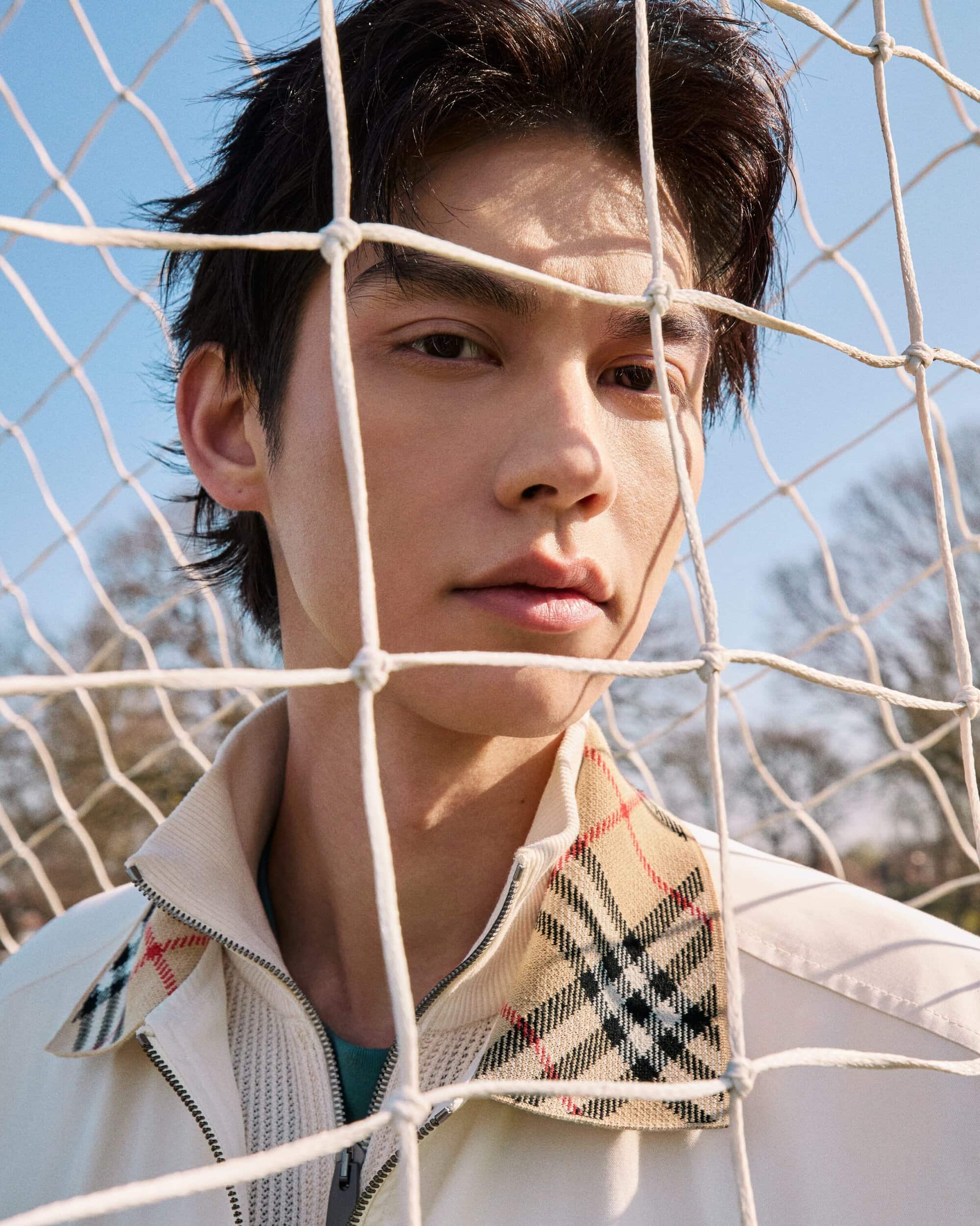 Bright photographed through a white goal net wearing a cream Burberry zip jacket with plaid collar detailing.