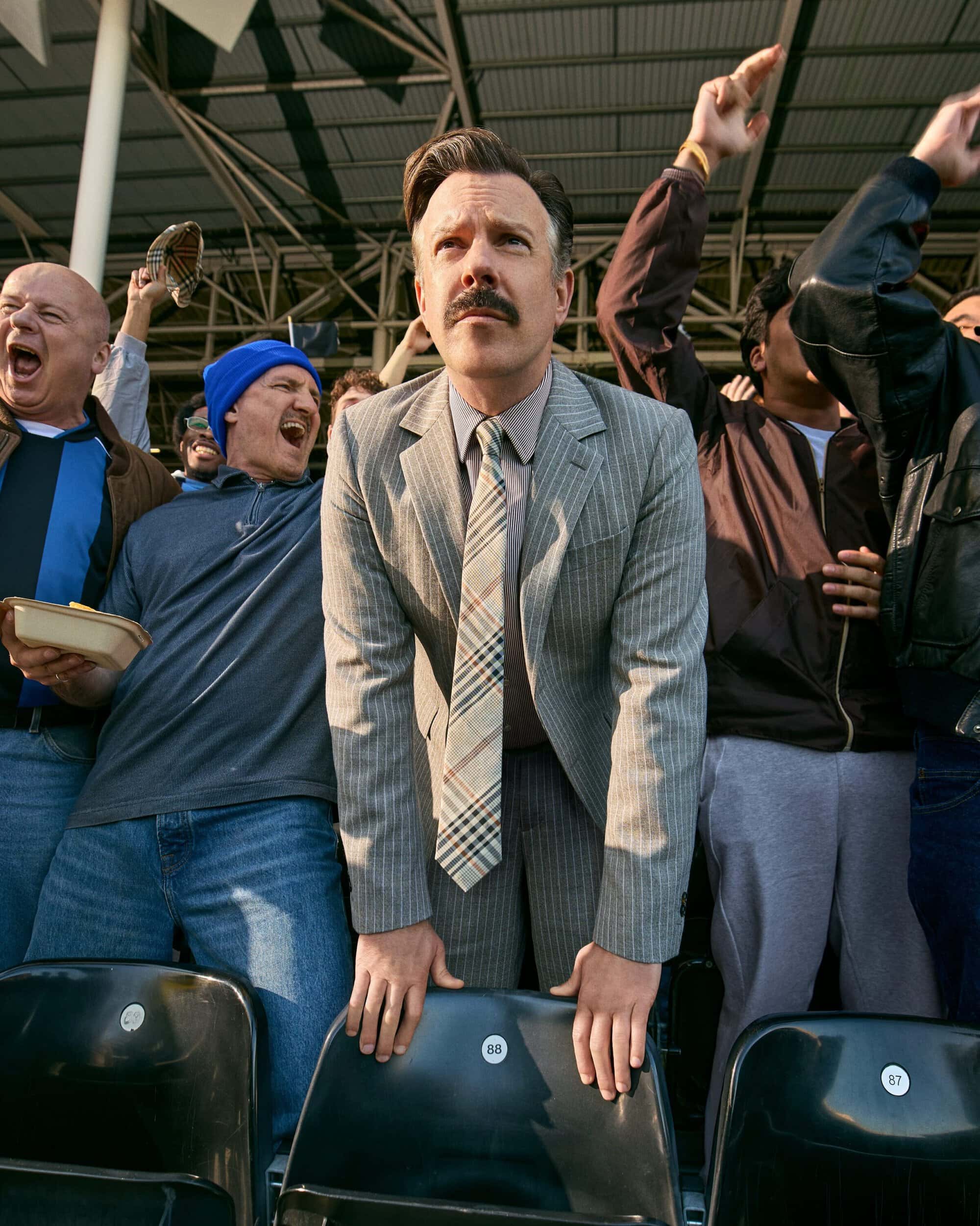 Jason Sudeikis wearing a grey pinstripe suit and plaid tie looking upward while standing among cheering sports fans in stadium seating.