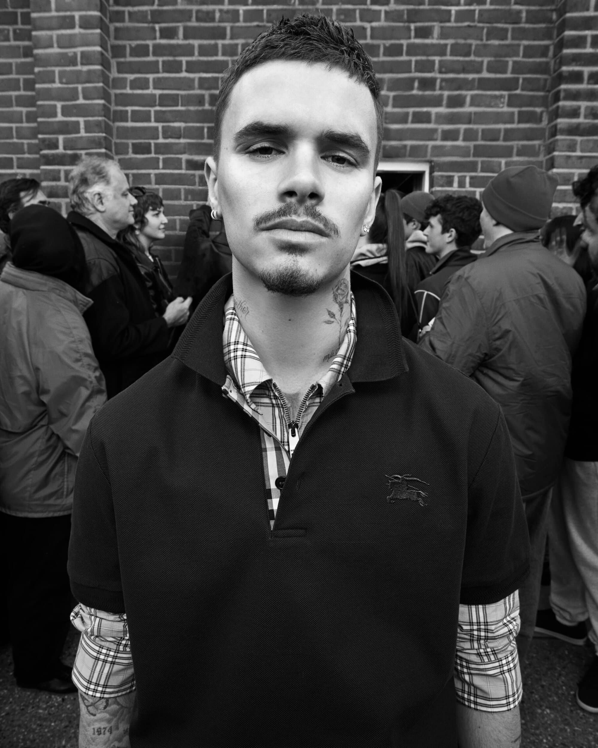 Black and white portrait of Romeo Beckham wearing a black polo shirt layered over a plaid collared shirt in front of a brick wall.