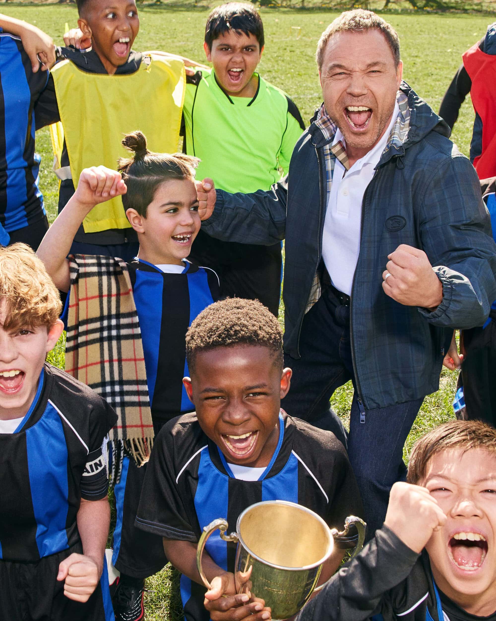 Stephen Graham and a youth soccer team cheering enthusiastically with a gold trophy on a grass pitch for Burberry.