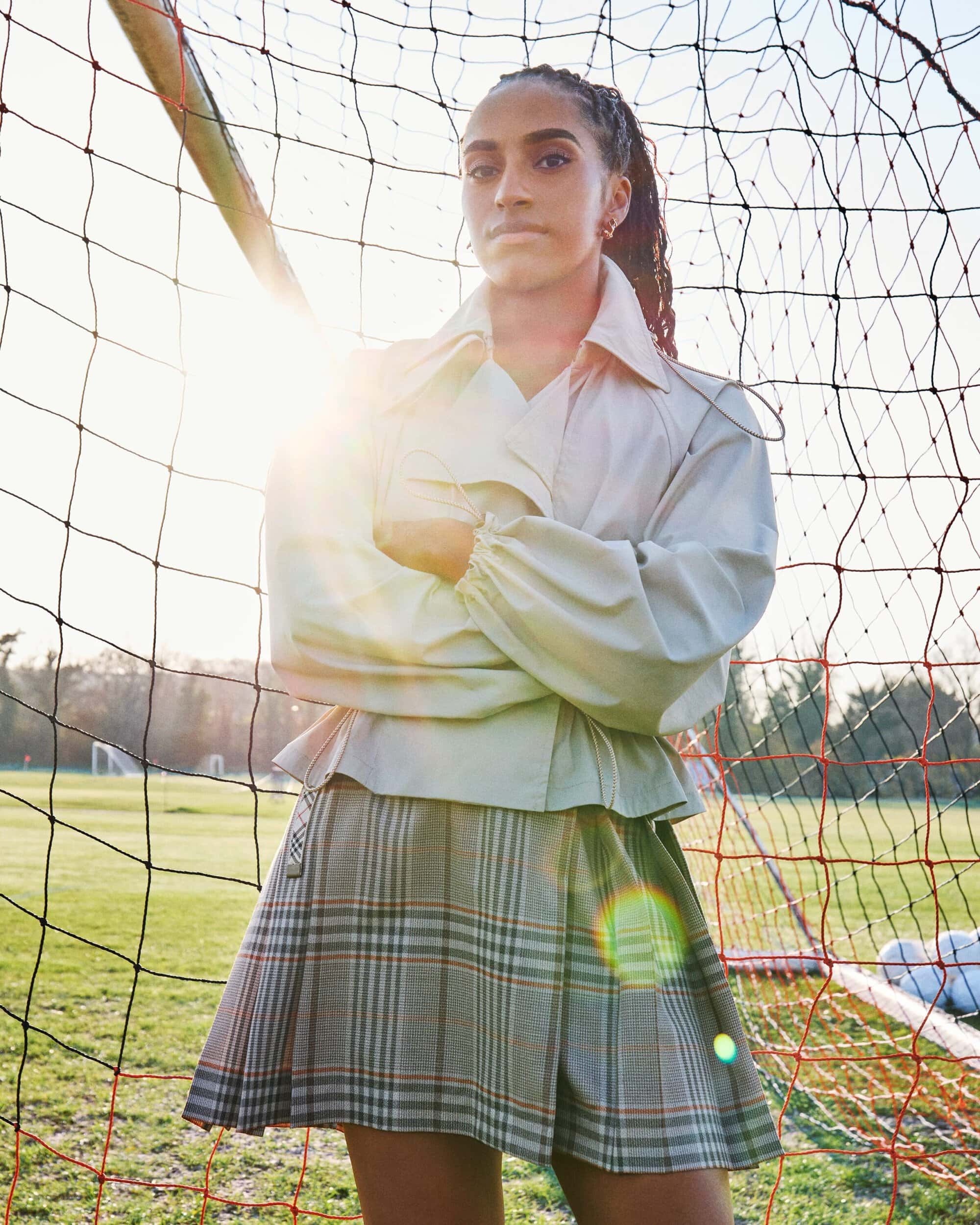 Leah Williamson standing with arms crossed in front of a soccer goal wearing a light jacket and plaid skirt