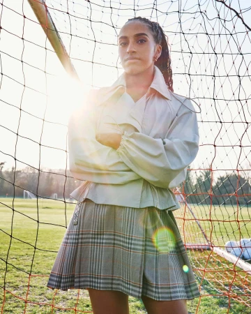 Leah Williamson standing with arms crossed in front of a soccer goal wearing a light jacket and plaid skirt