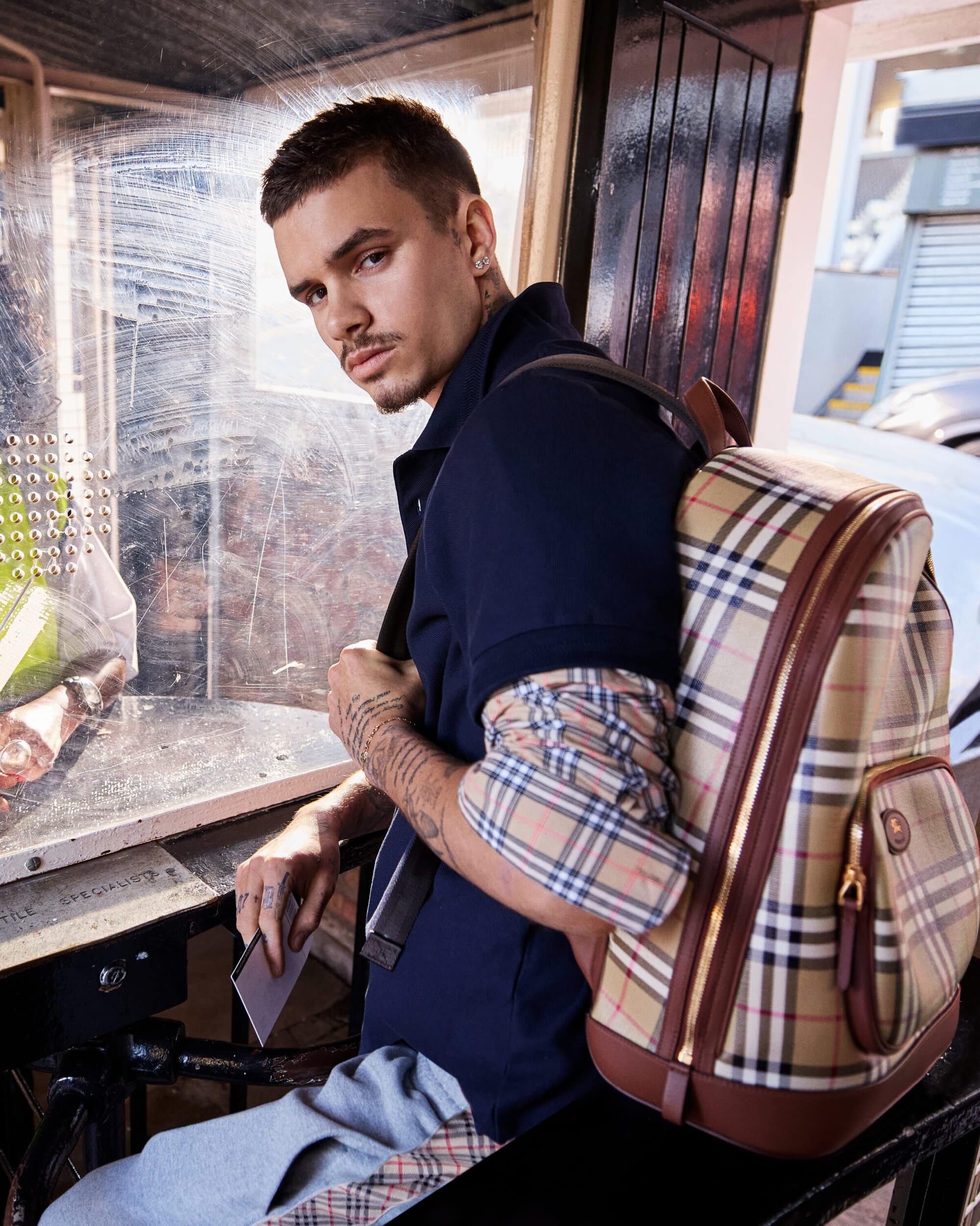 A stylish young adult male sits with a Burberry backpack in natural light, showcasing a blend of fashion and urban lifestyle.