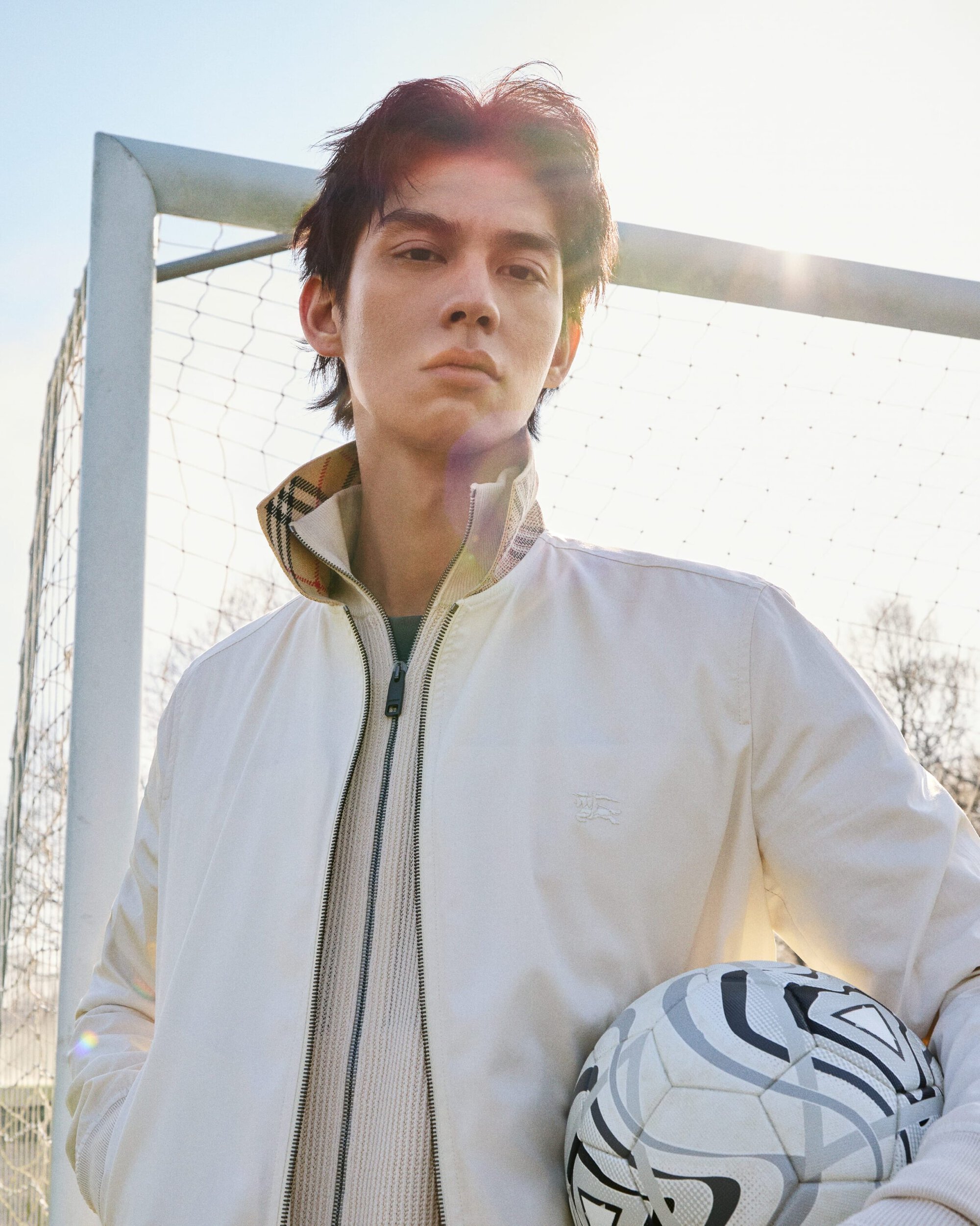 Bright Vachirawit holding a soccer ball in front of an outdoor goalpost wearing a cream Burberry zip jacket.