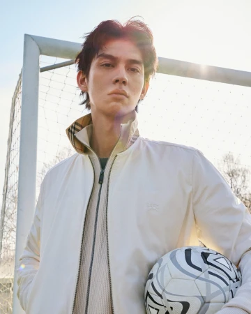 Bright Vachirawit holding a soccer ball in front of an outdoor goalpost wearing a cream Burberry zip jacket.