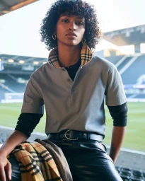 Model with curly hair in a grey polo shirt with check collar and black leather trousers seated in a sports stadium.
