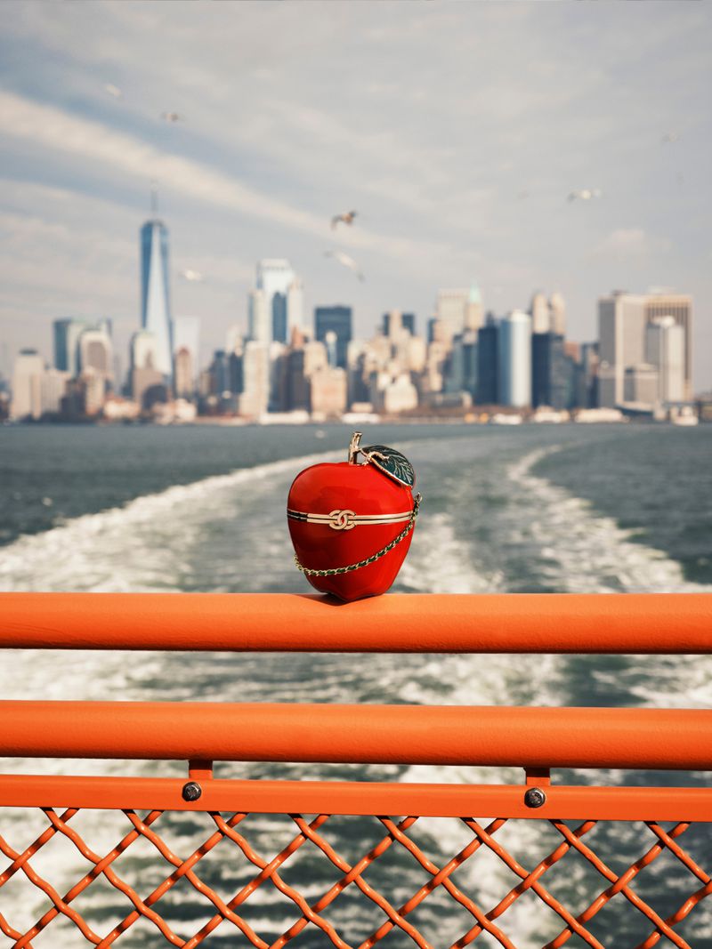 A red apple-shaped Chanel clutch with gold hardware sits on an orange ferry railing overlooking the New York City skyline.