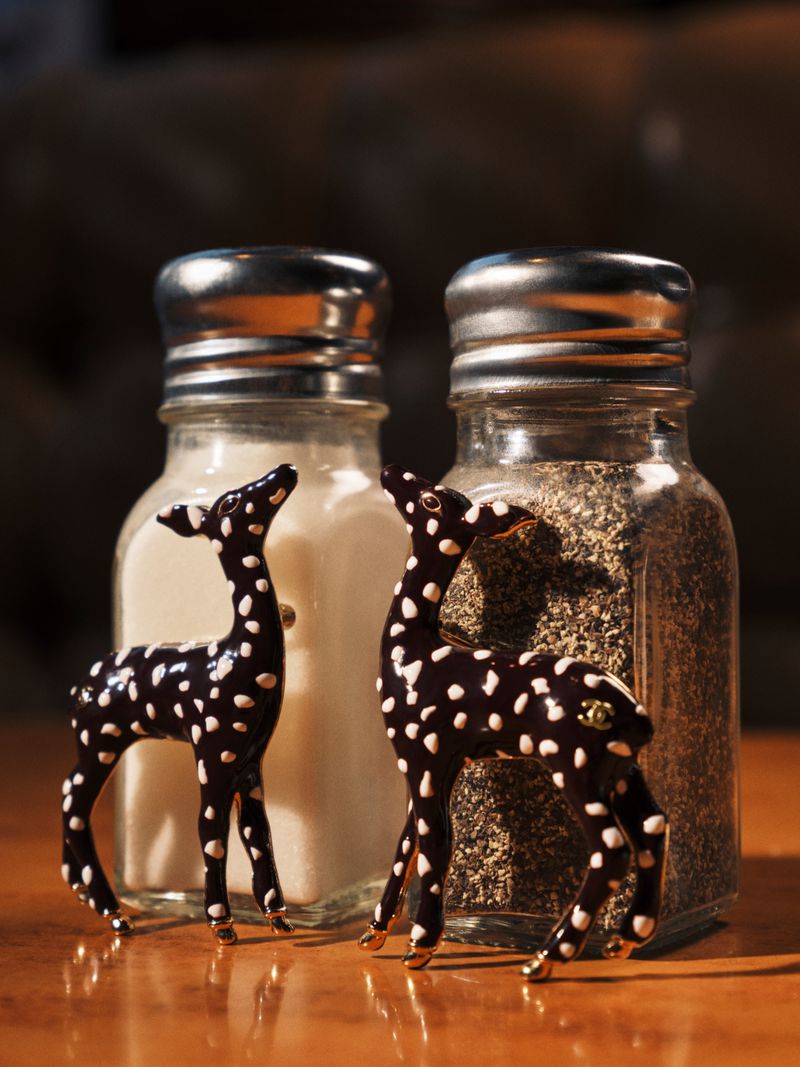 Two spotted fawn brooches standing next to glass salt and pepper shakers on a wooden tabletop.
