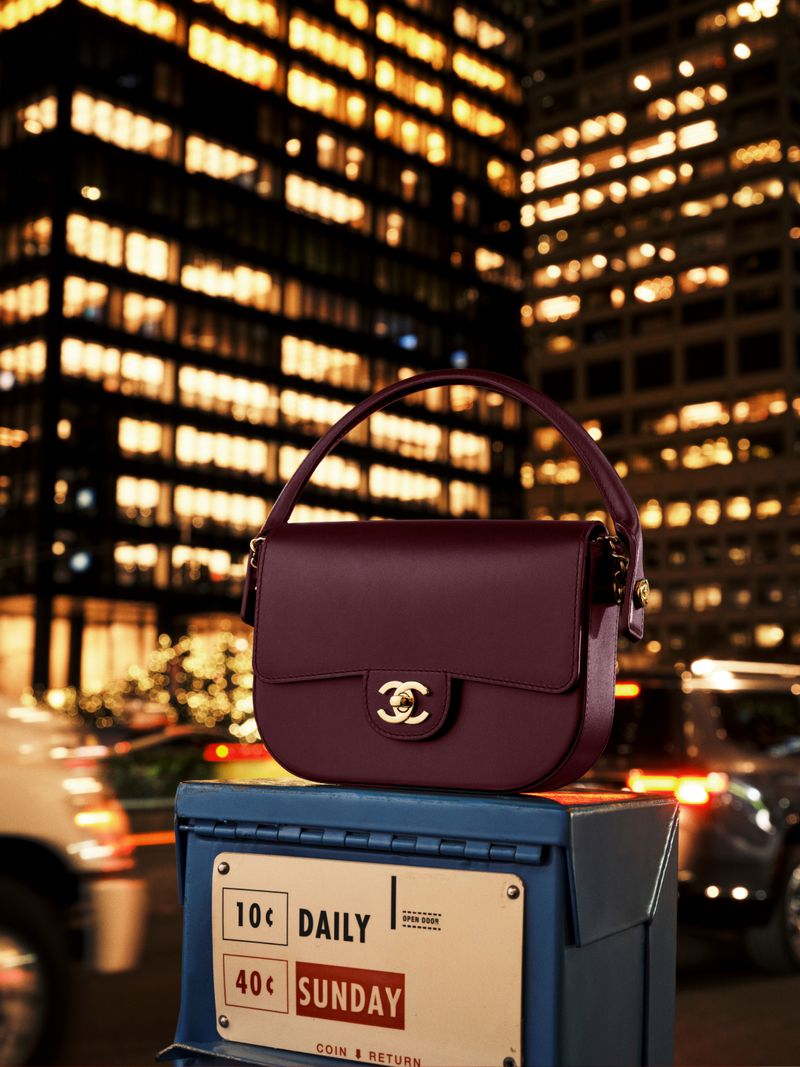 Burgundy Chanel handbag displayed atop a blue vintage newspaper dispenser against a blurred urban nightscape with illuminated buildings.