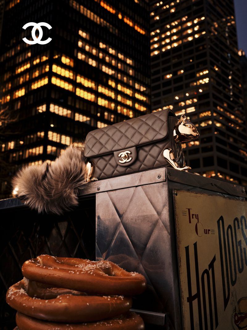 Chanel quilted handbag with sculpted metallic accents resting on a metal street food cart beside soft pretzels in front of city buildings at night.