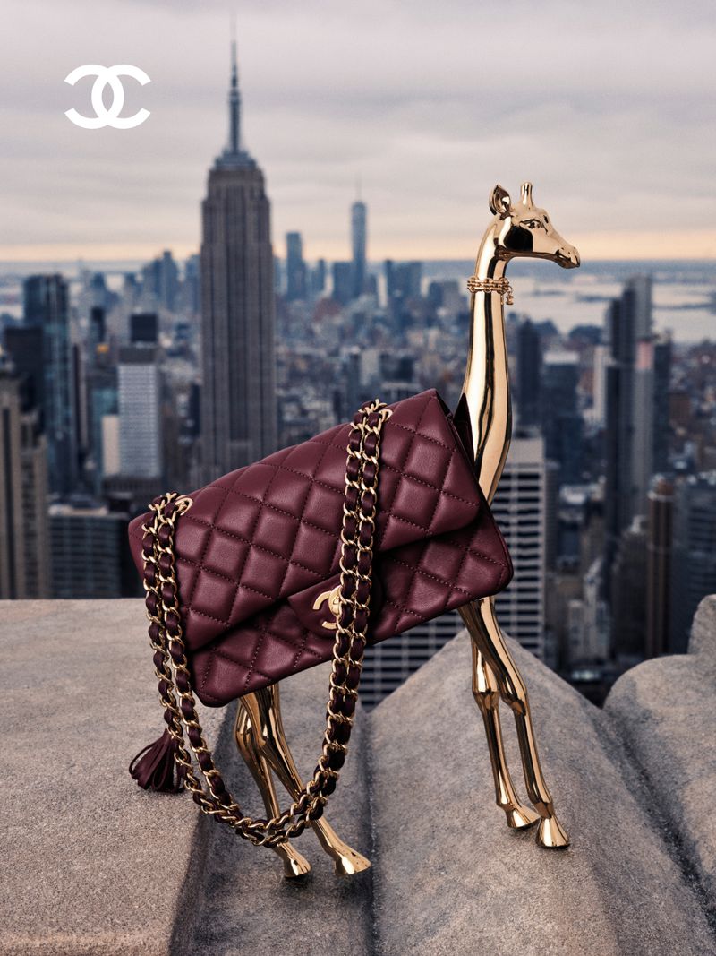 Chanel burgundy quilted leather handbag draped over a polished gold giraffe sculpture on a rooftop overlooking the New York skyline.