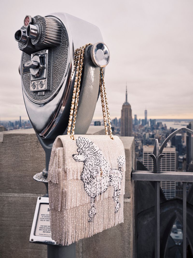 Embellished white fringed Chanel handbag with a beaded poodle design hanging on a coin-operated viewfinder overlooking a city skyline.