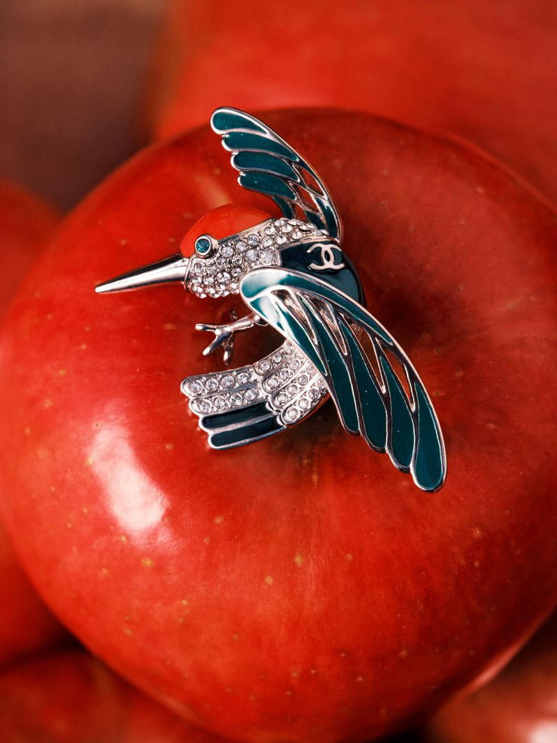 A Chanel bird brooch with teal enamel and pavé crystals resting on top of a red apple.