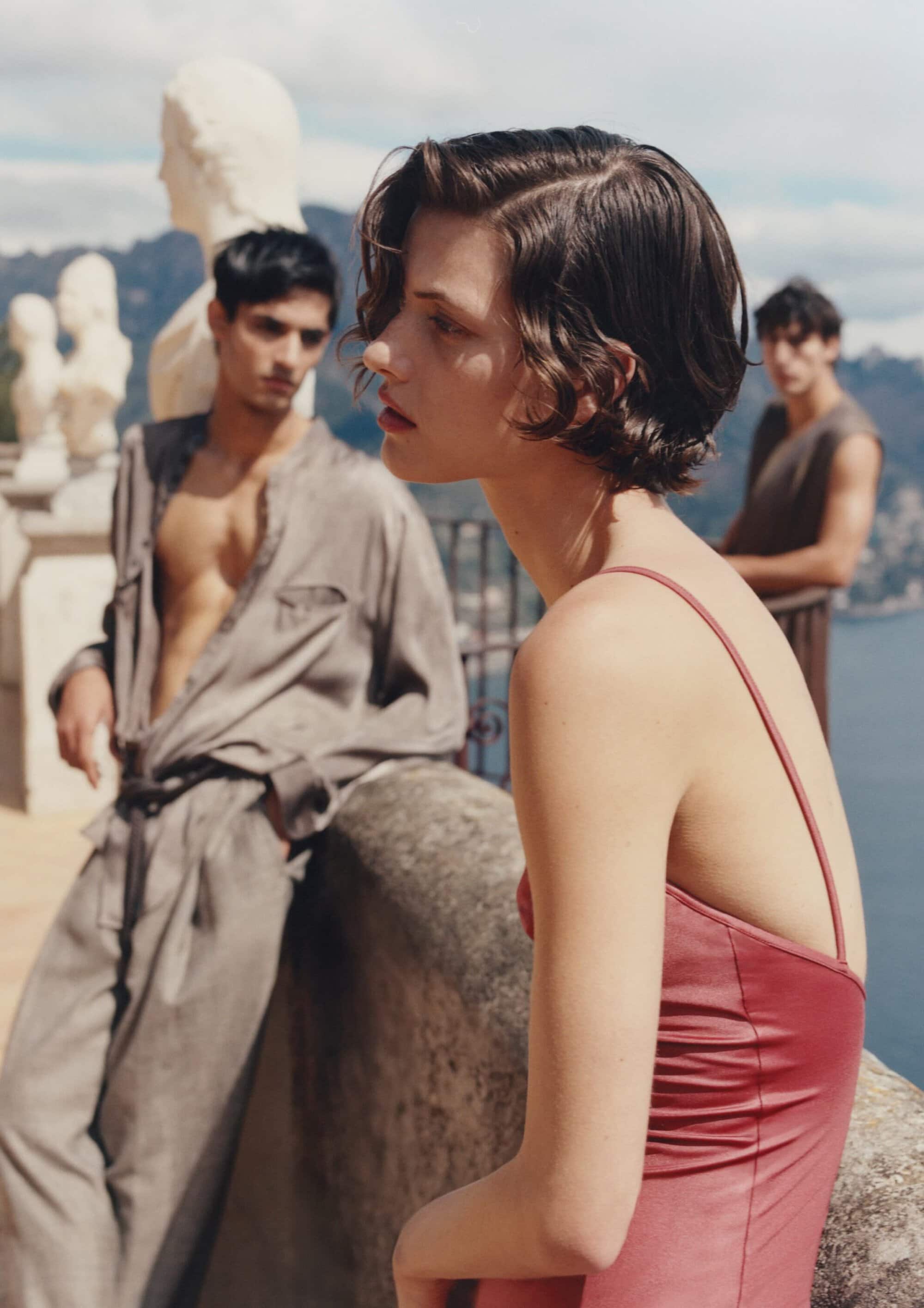 Woman in red dress in foreground with two men standing against coastal backdrop with classical statues