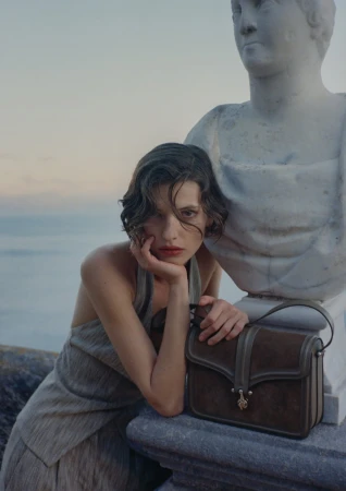 Valerie Scherzinger leaning beside a classical marble bust holding a brown suede Giorgio Armani handbag against a sea backdrop.