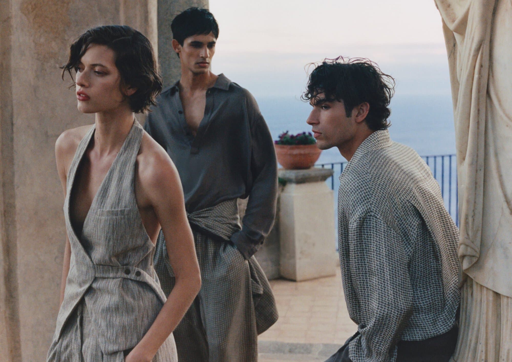 Model in a grey tailored halter vest posed outdoors on a stone terrace with two male models overlooking the sea.