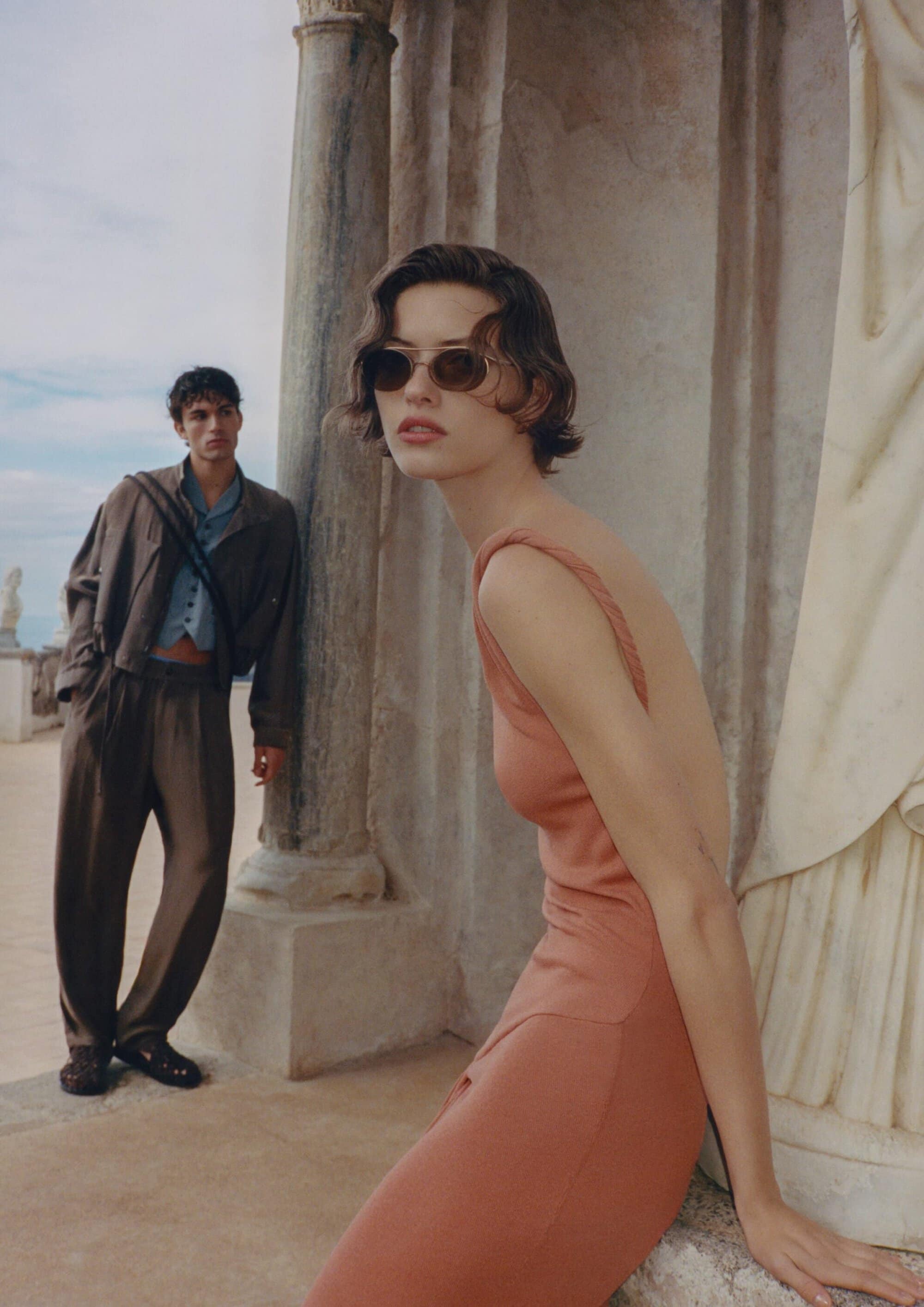 Model Valerie Scherzinger in a coral backless dress and sunglasses with Balthazar Dib standing near stone pillars.