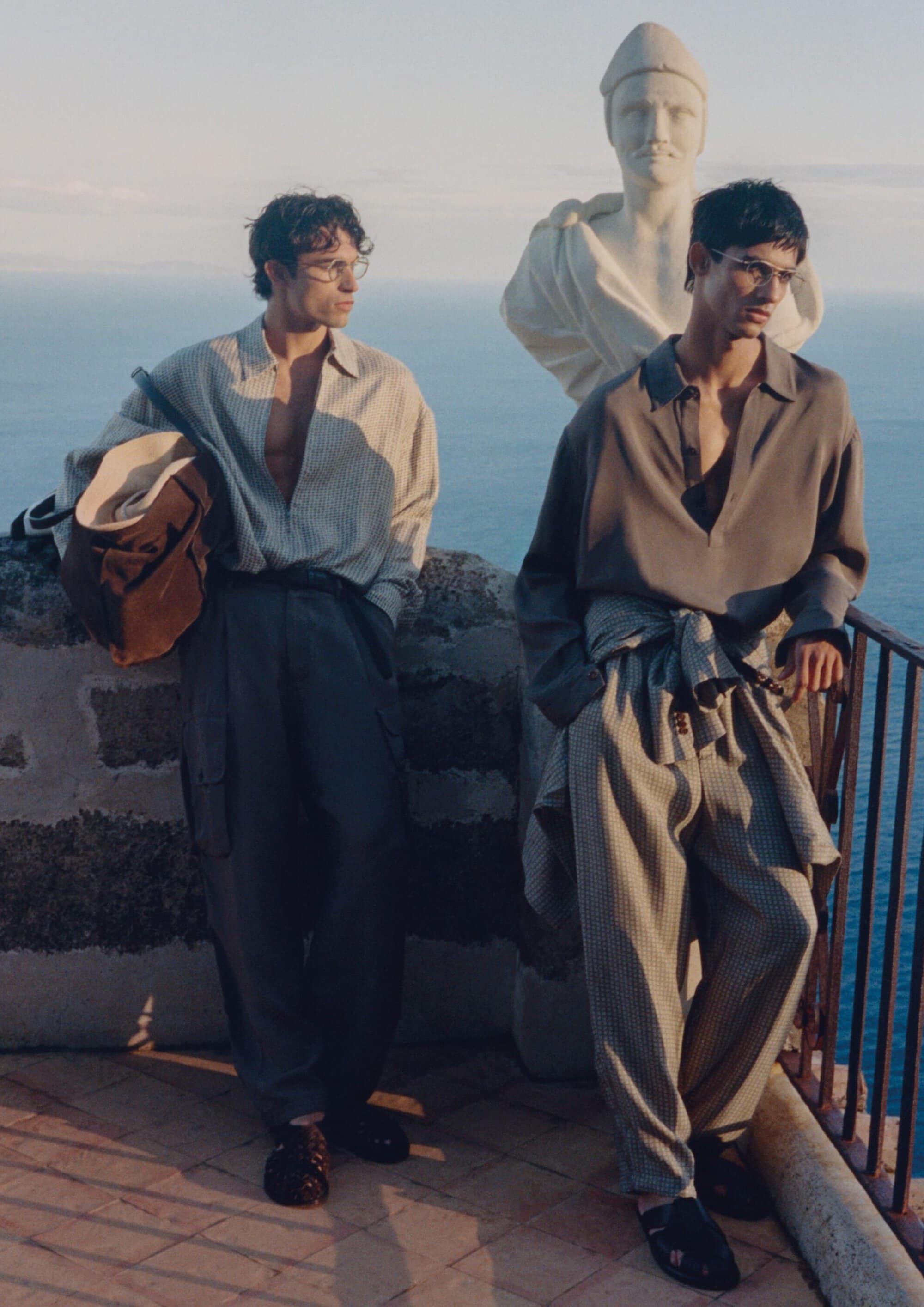 Two male models in relaxed neutral menswear pose beside a classical marble bust on a scenic cliffside terrace overlooking the ocean.