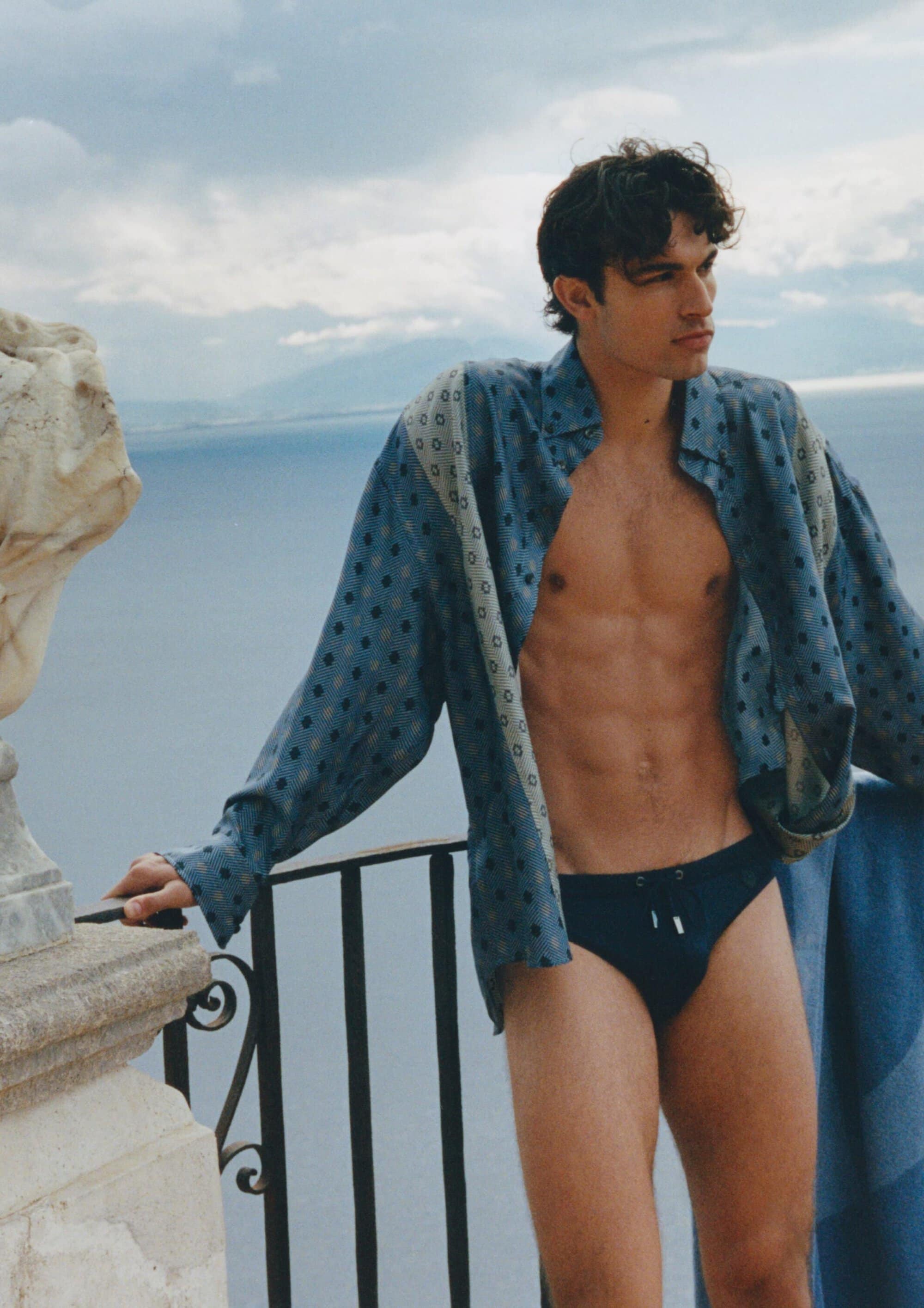 Man in an unbuttoned blue printed shirt and dark swim briefs leans against a balcony railing overlooking the sea.